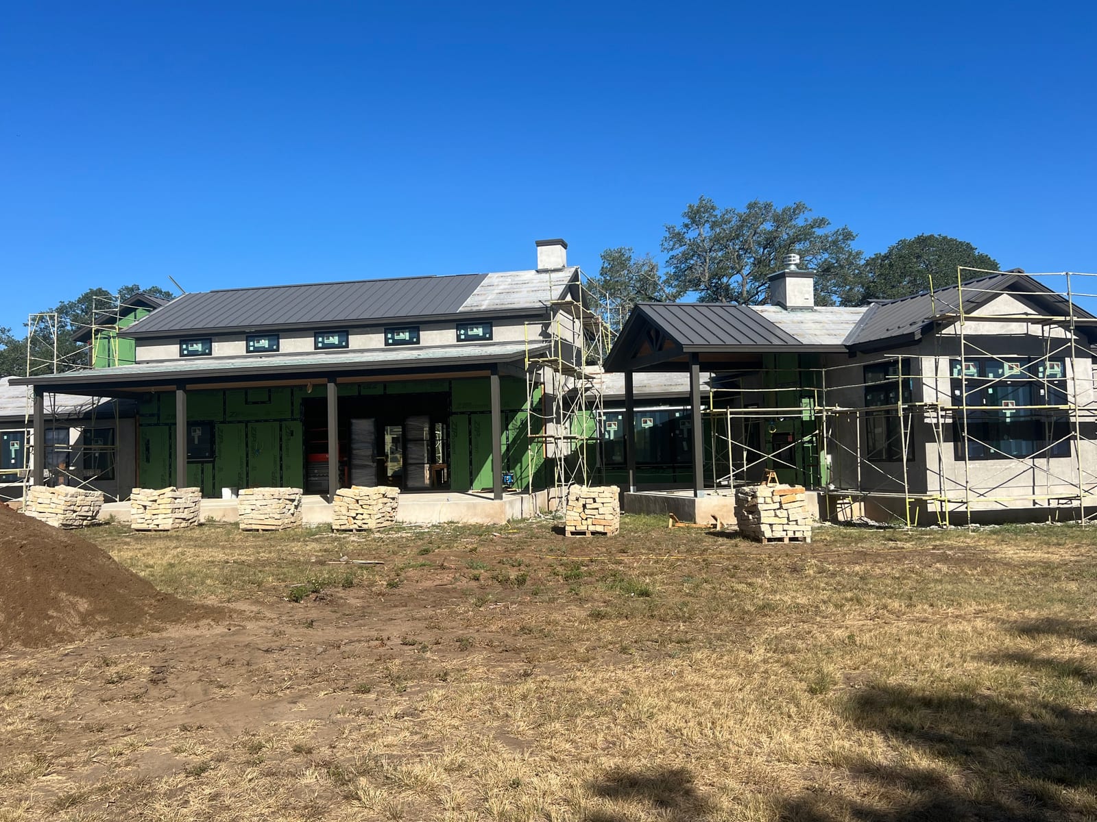 Long elevation of a new Hill Country home with a dark standing seam roof over a deep covered porch and a stone chimney through the ridge