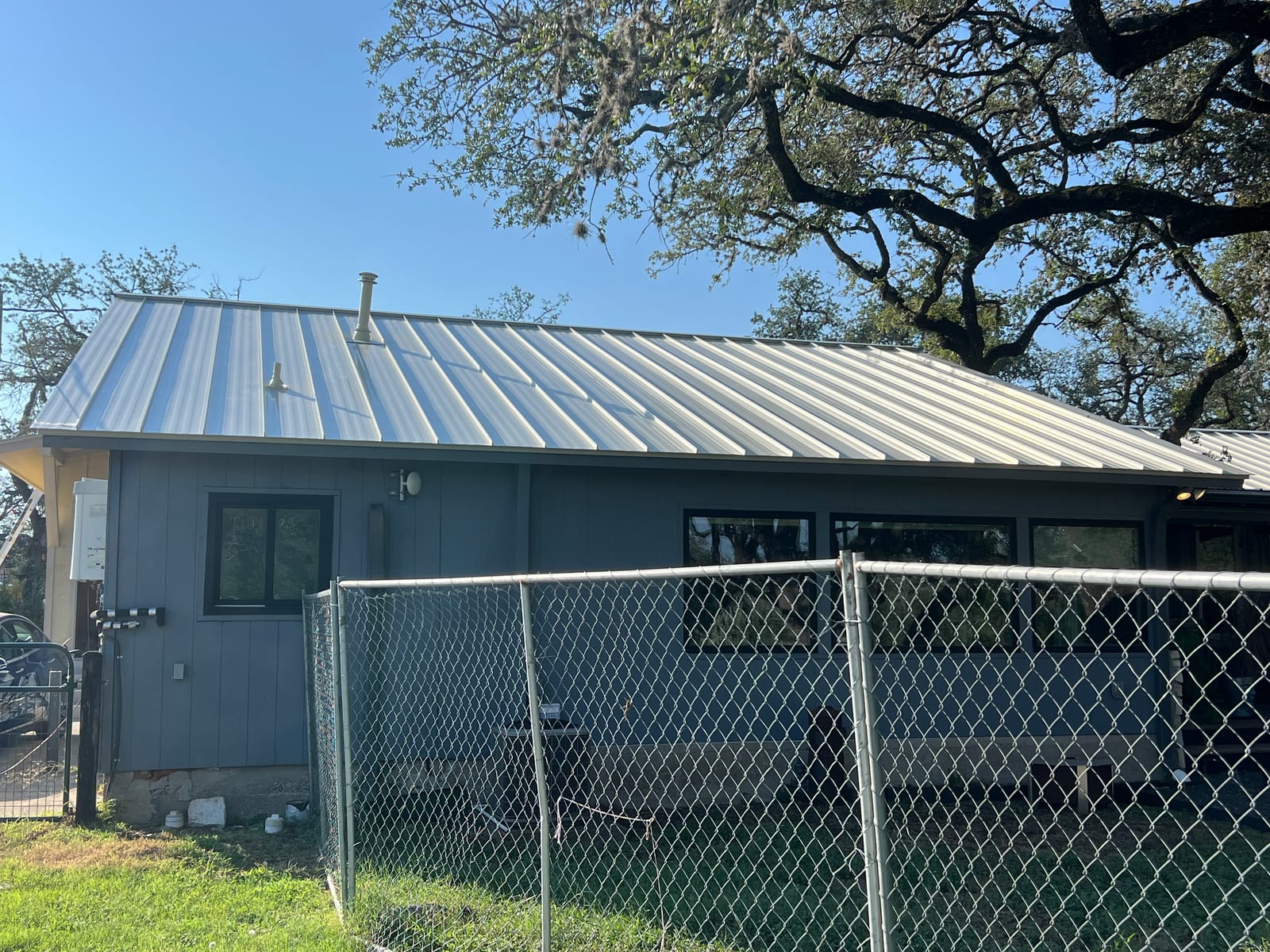 Completed sandstone metal roof over a grey board-and-batten wing of the house, with a flue and pipe boot set into the panels and the ridge closed
