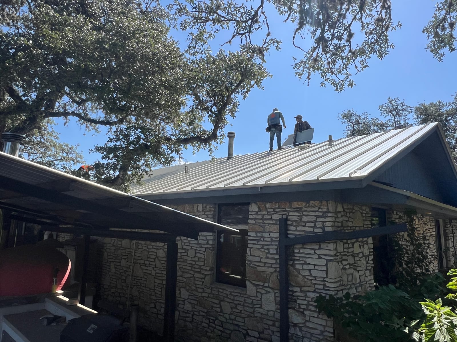 Two roofers standing on the ridge of a partly finished metal roof above a stone house, carrying a panel into place