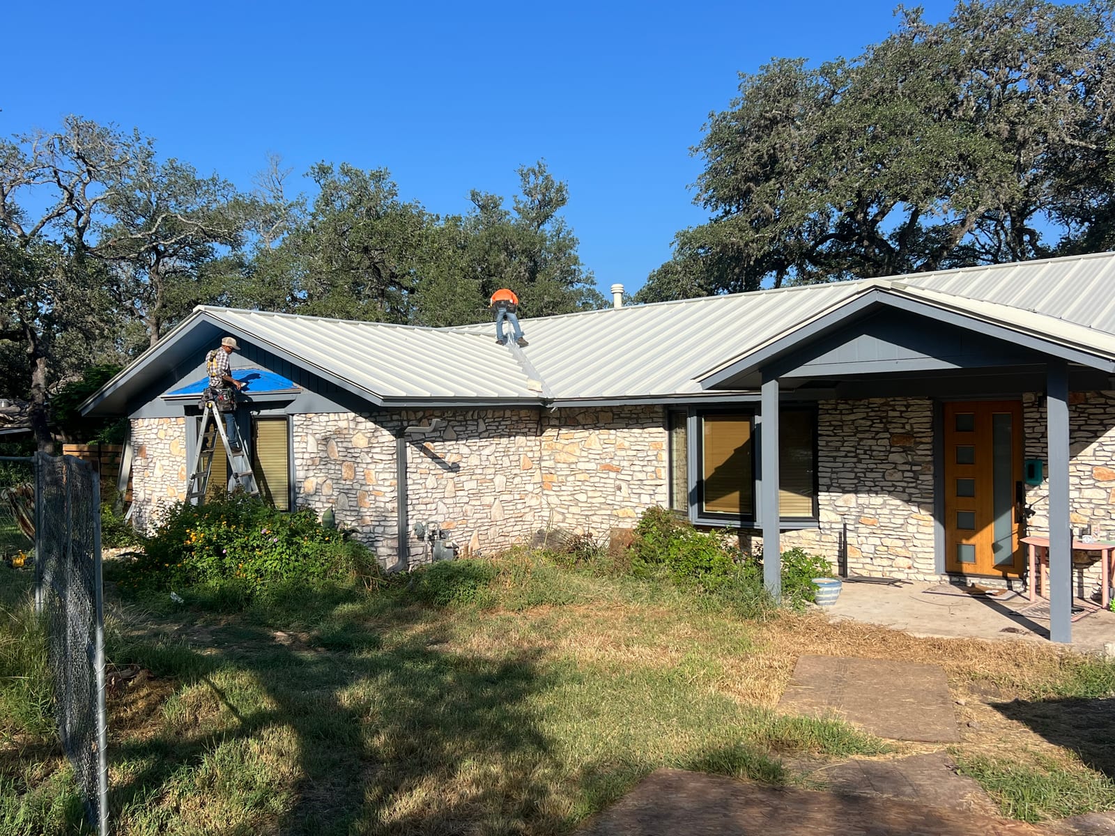 Roofers working on a metal roof over a stone Austin house, one on a ladder at the gable end and one on the ridge, with live oaks behind