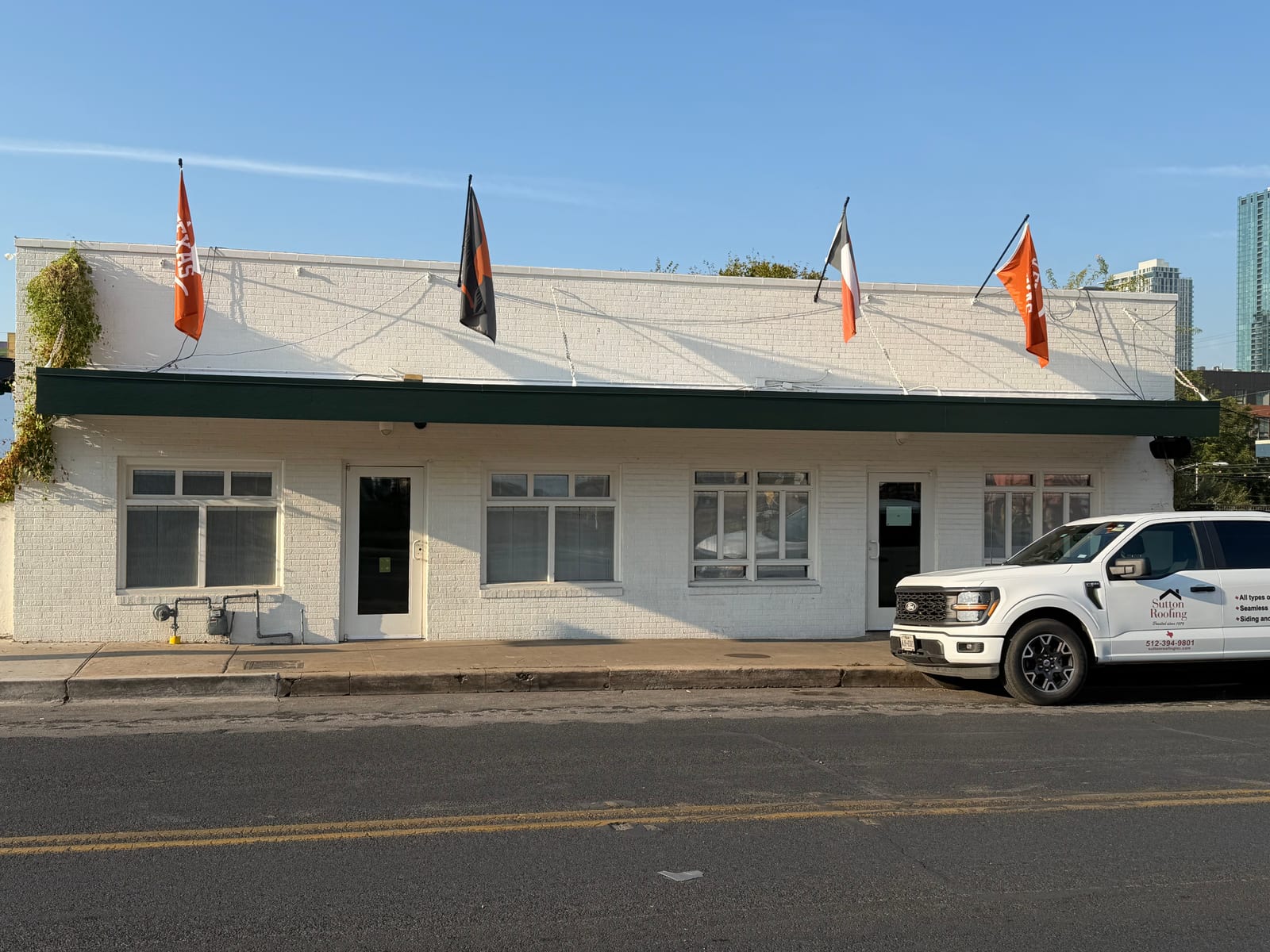 White brick building on East 6th Street with a dark green fascia along the canopy roof and a Sutton Roofing truck parked out front