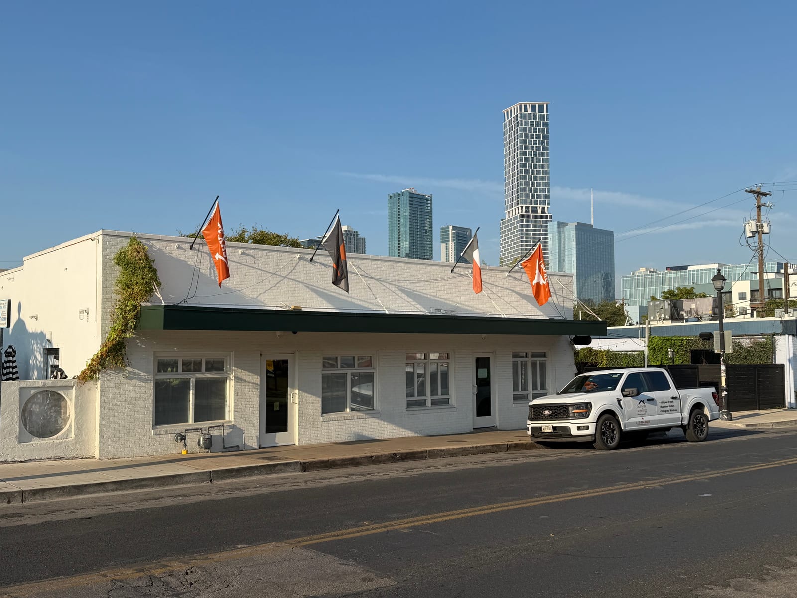 Single-story white brick building with a green canopy fascia and the downtown Austin skyline behind it