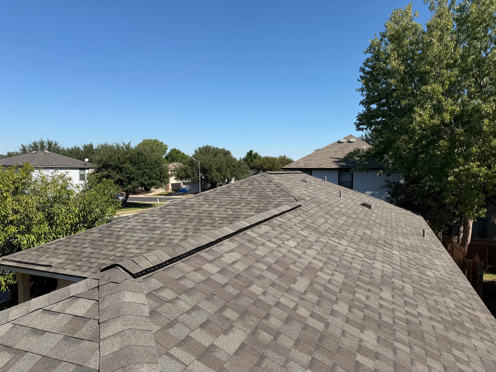 New shingle roof with a continuous ridge vent running the length of the main ridge, neighboring rooftops and trees beyond