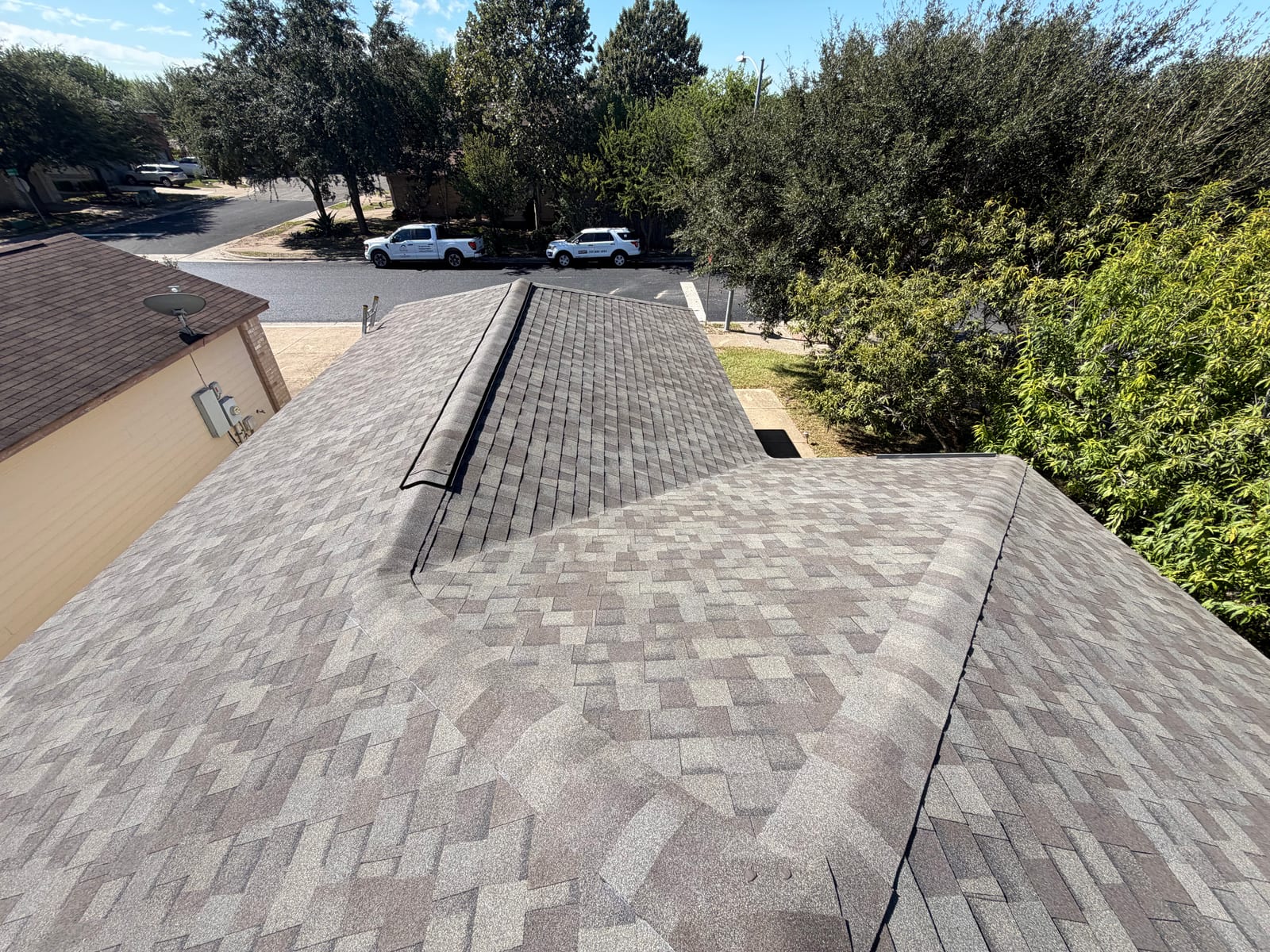Looking down the front hip of a new grey-brown shingle roof toward the street, with Sutton Roofing trucks parked below